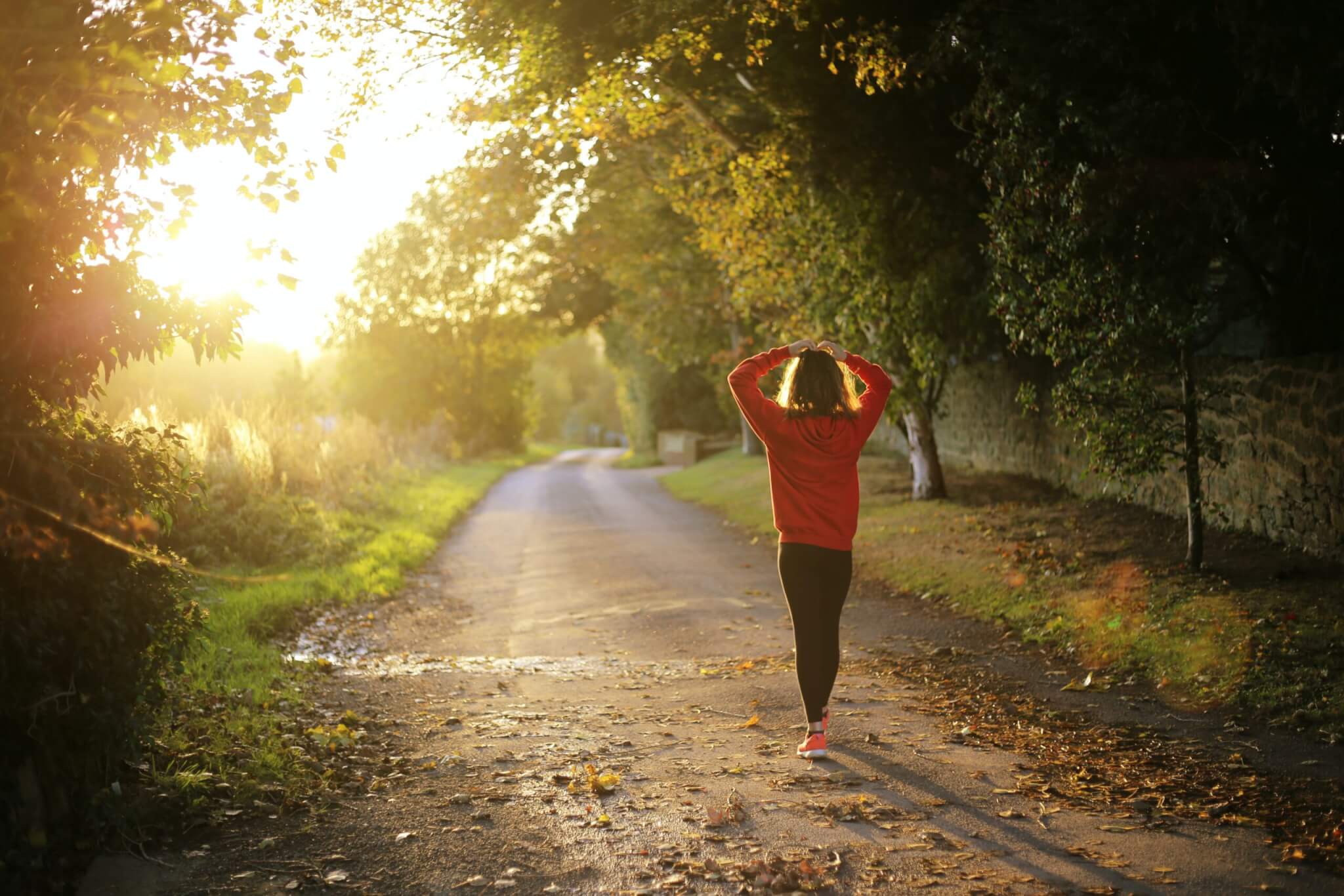 A person in a red sweatshirt walks alone down a sunlit, tree-lined country road, hands resting on their head. Autumn leaves cover the ground as sunlight filters through the trees—suggesting a moment when one might wonder: How Can Earthing Affect Stress?.