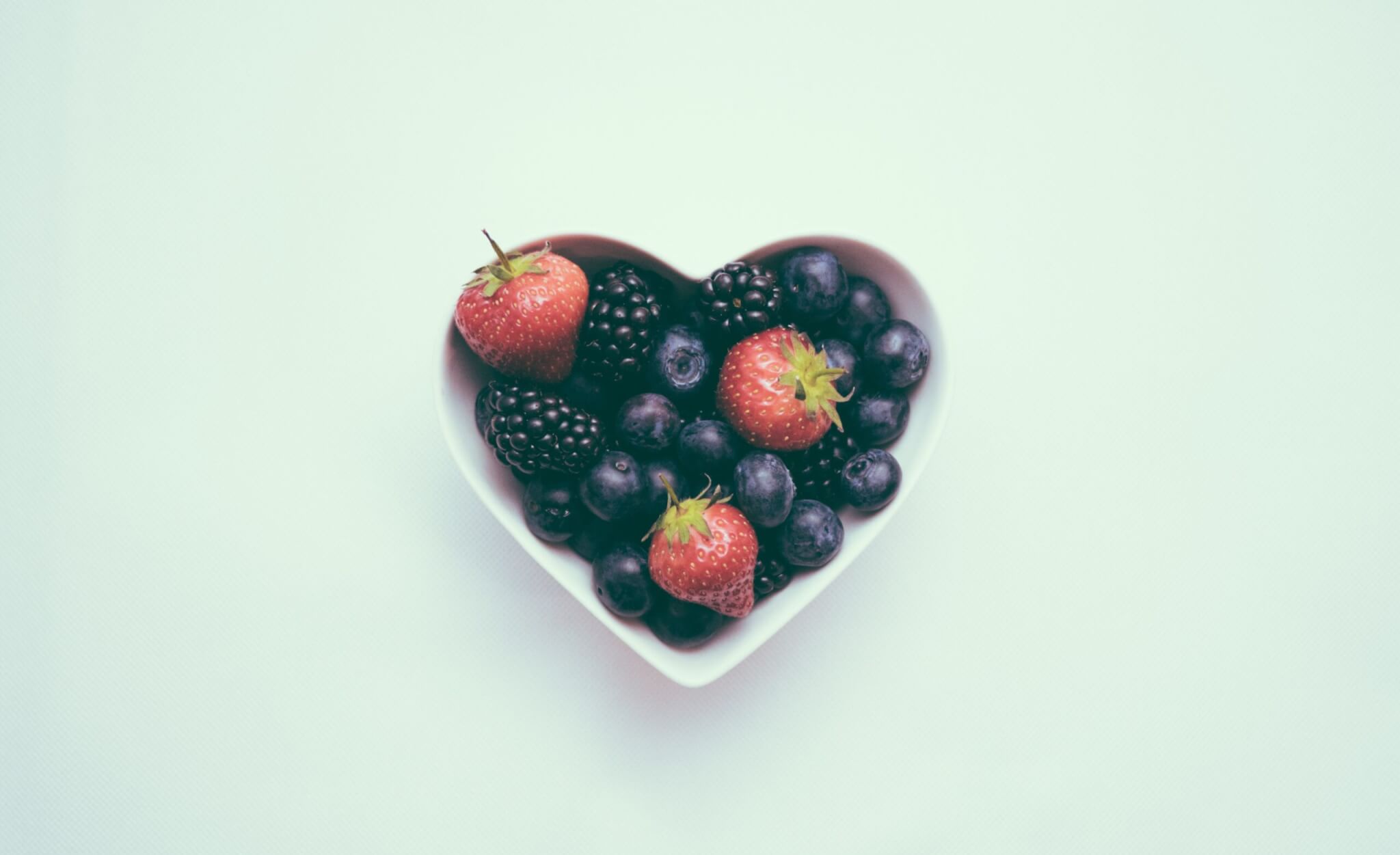 A white, heart-shaped bowl filled with strawberries, blueberries, and blackberries sits on a light background—perfect for anyone interested in how to boost heart health through grounding and nutritious foods.
