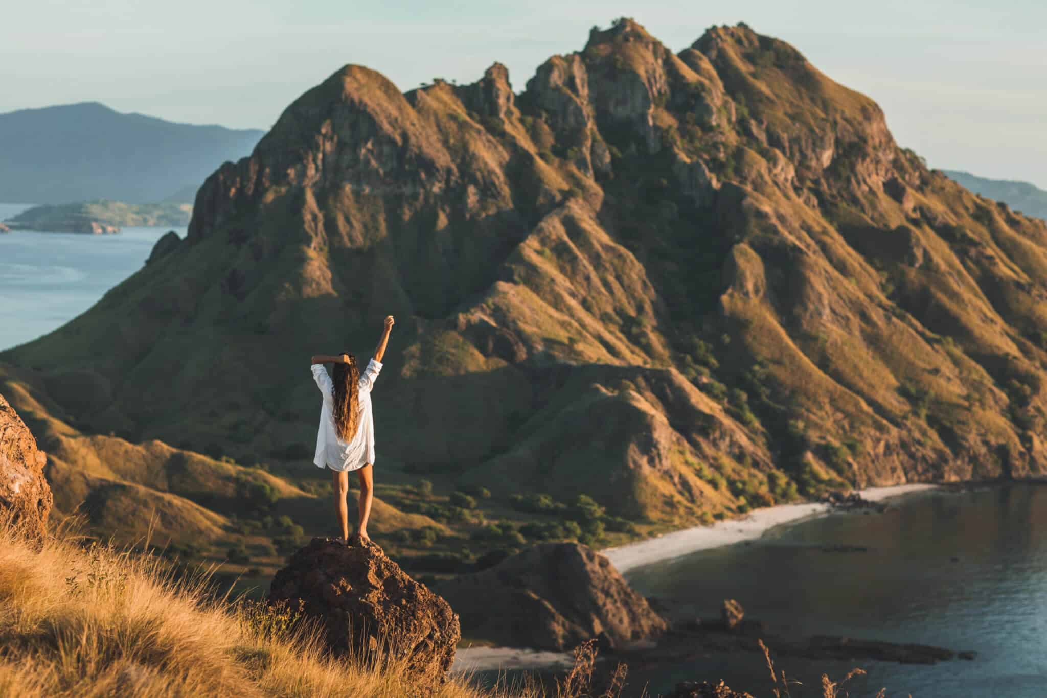 A person stands BeGrounded on a rock with one arm raised, facing a vast, rugged mountain and coastline, surrounded by grassy hills and shimmering water beneath a clear sky.