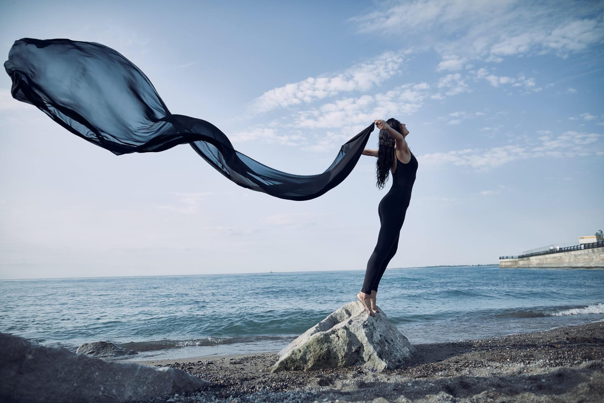 A woman in a black outfit stands barefoot on a rock by the sea, grounded on the rugged surface as she holds a long, flowing black fabric that billows dramatically in the wind under a partly cloudy sky. What surfaces can I be grounded on?.