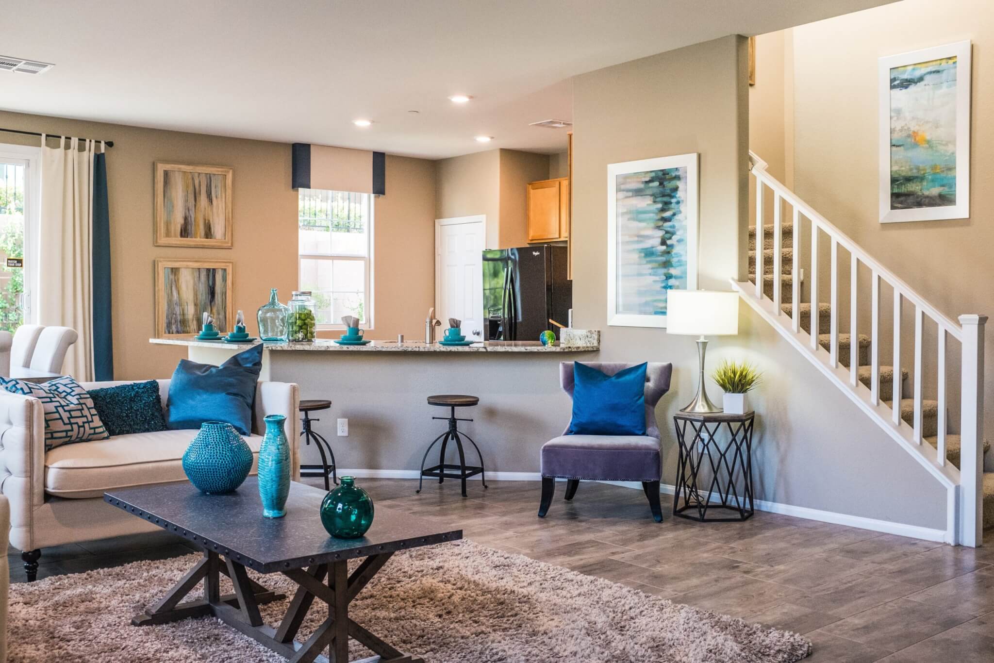 Modern living room with neutral walls, blue accents, a white sofa, armchair, coffee table with vases—showing you can be grounded indoors surrounded by inviting bar stools at the kitchen counter, wall art, lamp, and carpeted stairs with a railing leading upward.