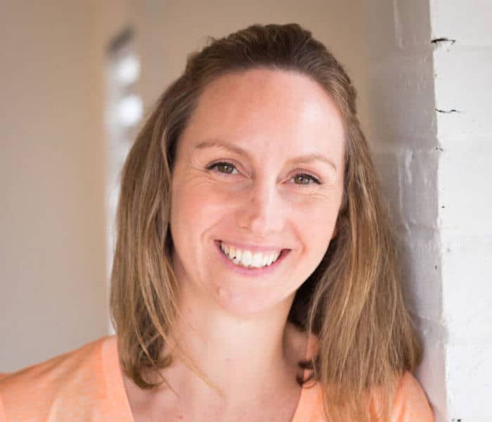A woman with shoulder-length light brown hair smiles warmly while leaning against a white brick wall. She is wearing a light orange top in a softly lit indoor setting, reflecting the welcoming spirit of About BeGrounded.