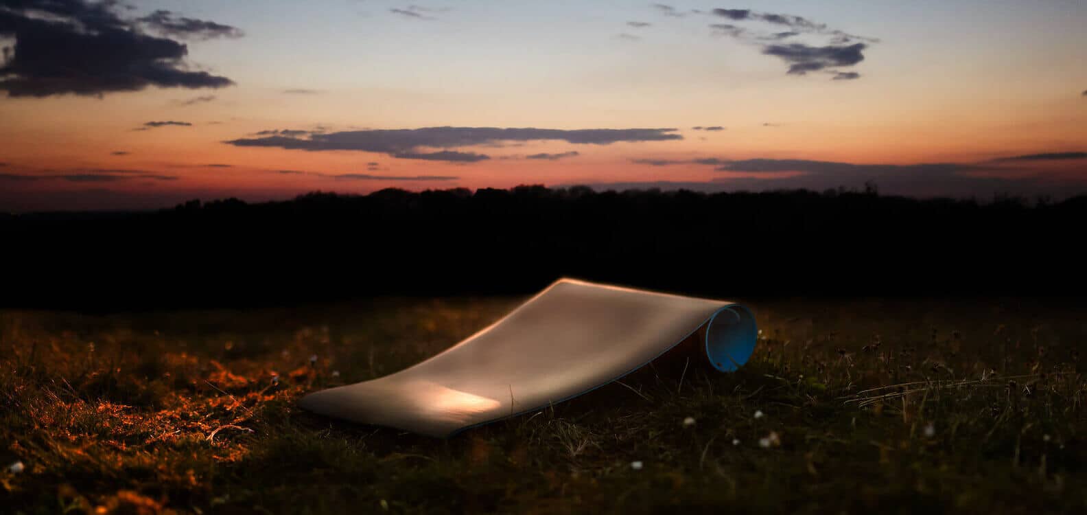 A modern, curved BeGrounded lounge chair sits alone on grass at dusk, softly illuminated, with a dark forest and a colorful sunset sky in the background.