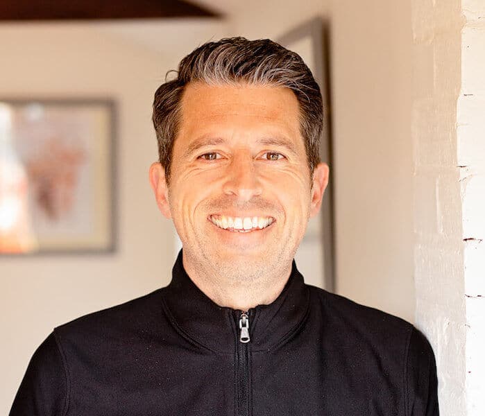 A man with short dark hair, wearing a black zip-up jacket, smiles while standing indoors near a white wall, with blurred artwork visible in the background—capturing the calm, approachable spirit of About BeGrounded.