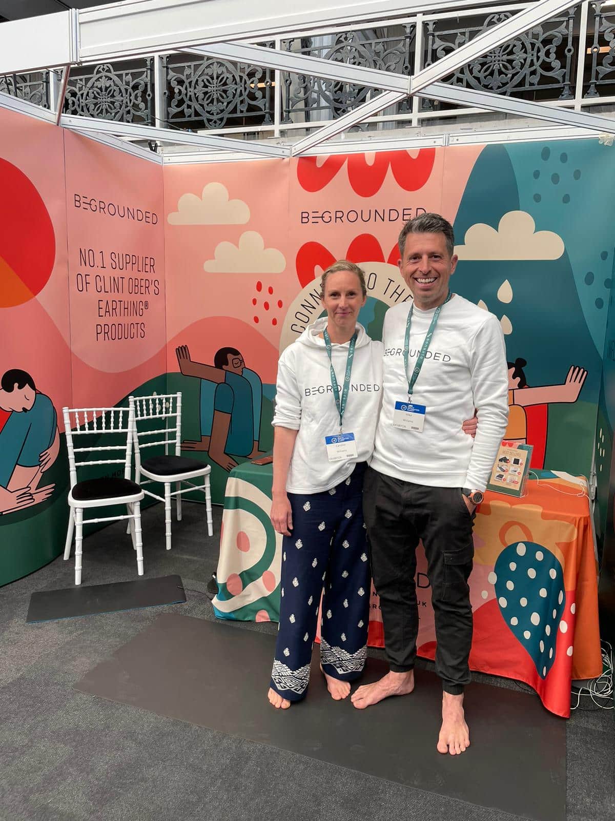 Two people stand barefoot in a colorful booth with “BEGROUNDED” branding. They wear matching white shirts and name badges, smiling at the camera. The booth displays products and illustrated banners in the background.