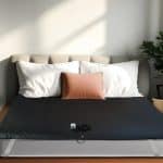 A neatly made bed with white pillows, a tan accent pillow, and the Earthing® Grounding Mattress Cover - Super King Size featuring a small electronic device and cord; sunlight streams in as a green potted plant sits nearby.