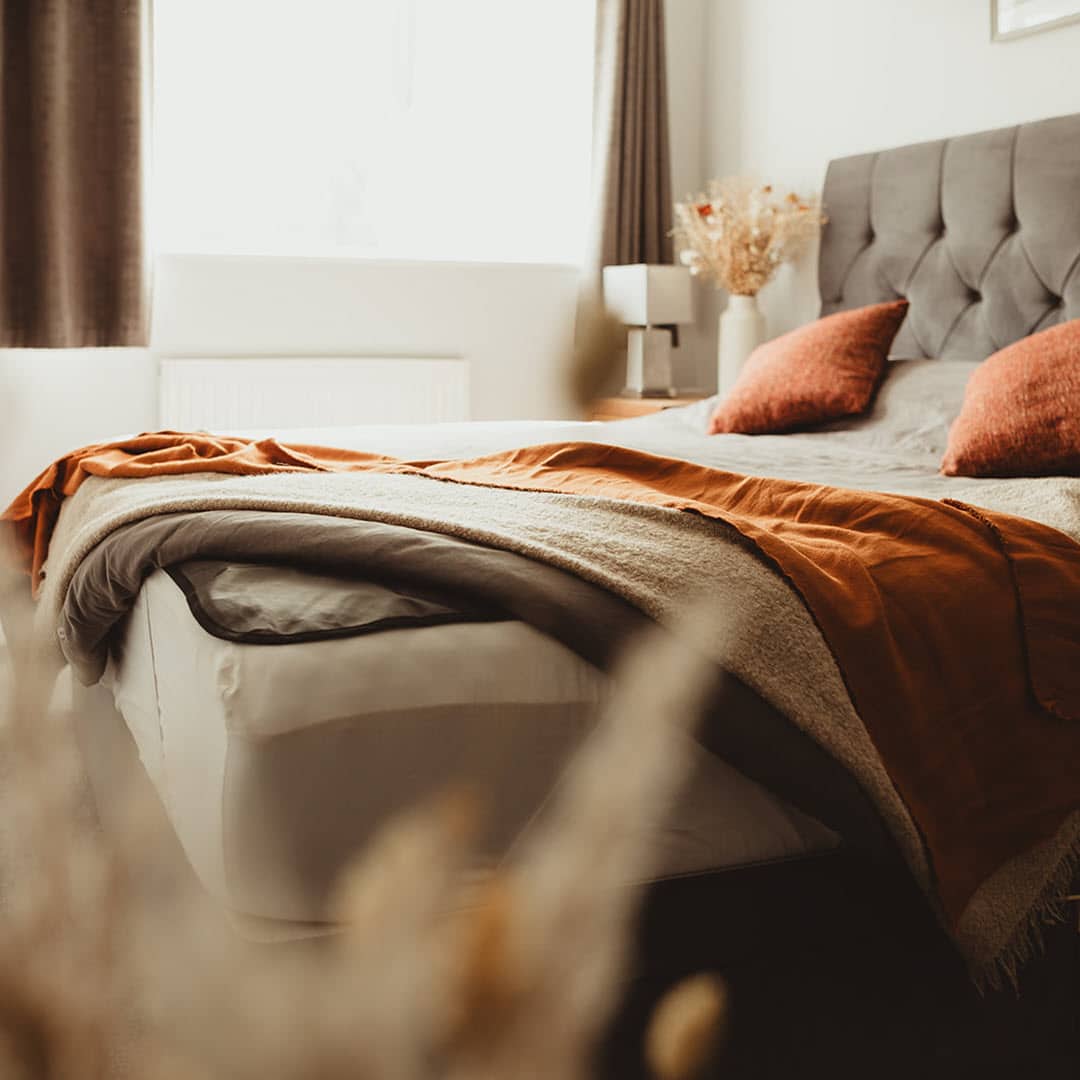 Cozy bedroom with a neatly made bed featuring gray sheets, an orange blanket, and two orange pillows. A tufted headboard is visible, and sunlight streams in from a window with dark curtains. A small side table holds a lamp and dried flowers.