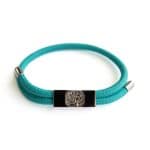 The Earthband PEMF Bracelet features a turquoise cord, silver metal tips, and a rectangular black charm with a silver tree design, inspired by PEMF wellness and shown on a white background.