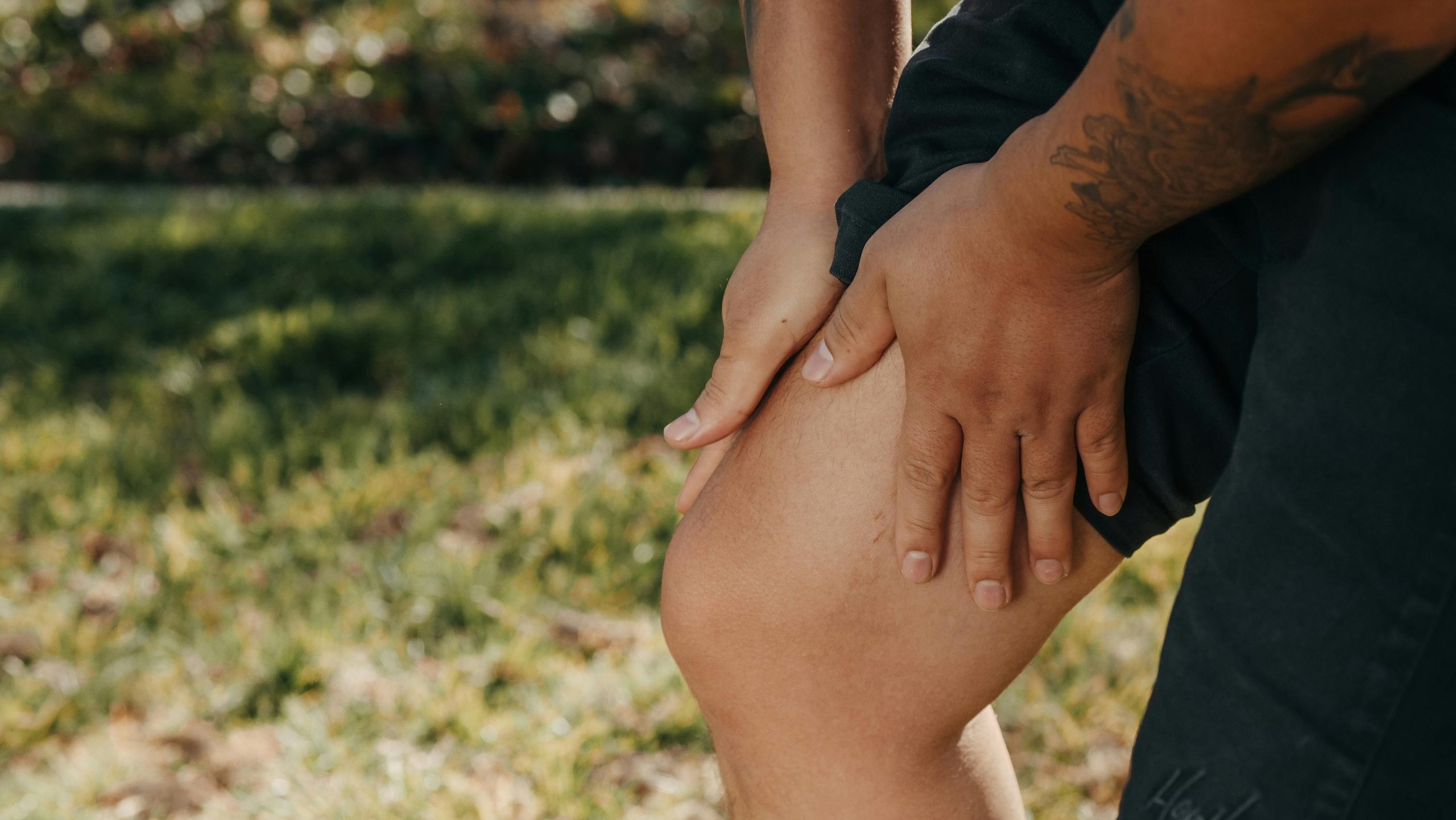 A person outdoors wearing black shorts holds their knee with both hands, appearing to experience pain or discomfort. Practicing anxiety grounding exercises could help manage stress in this calming setting with grass and foliage in the background.