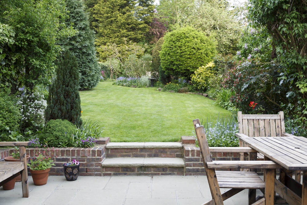 A well-kept backyard garden with a lawn, lush green trees, and flower beds offers the perfect spot for earthing at home. A wooden patio table with chairs sits on a stone terrace, while potted plants line the brick wall beside the steps to the grass.